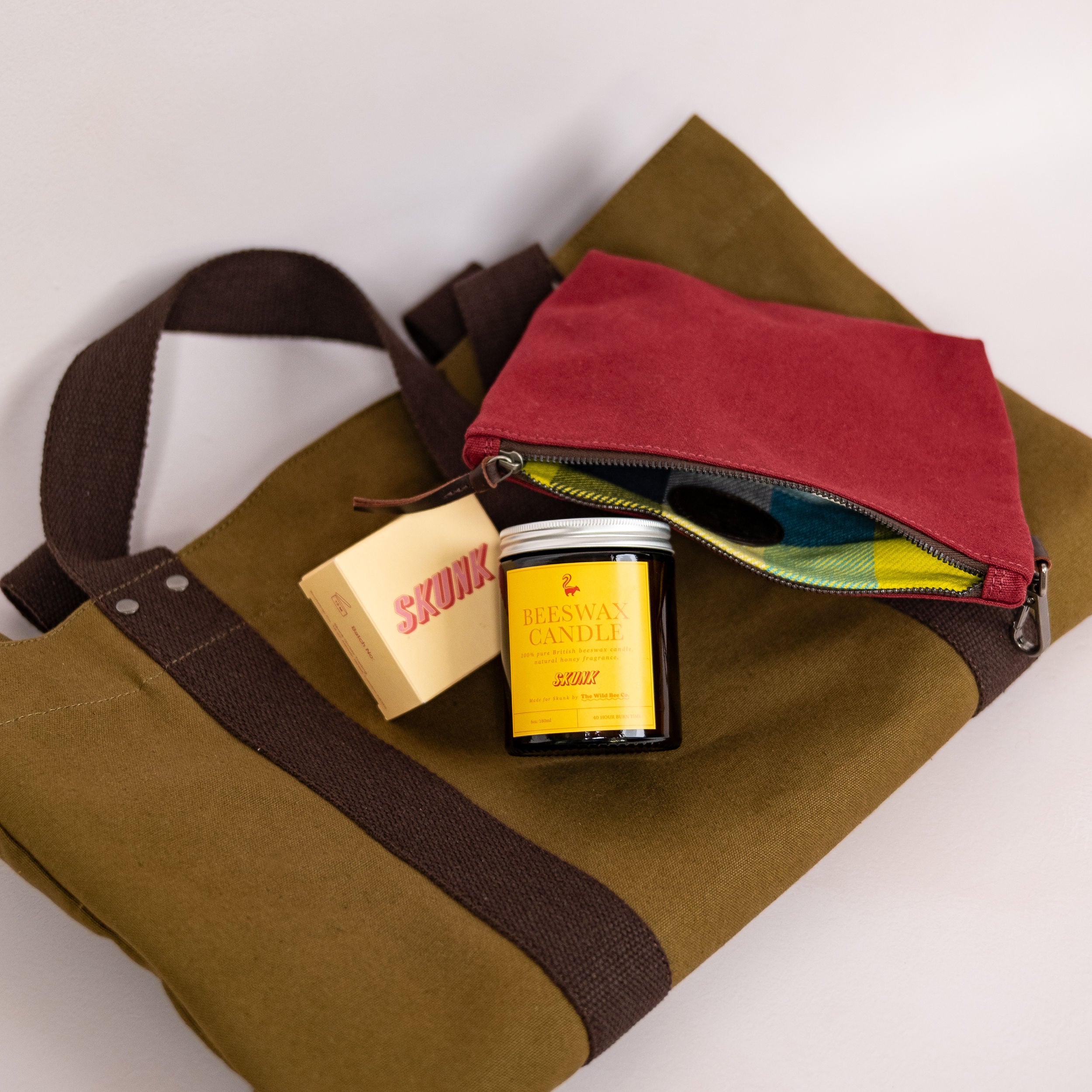 Skunk's olive green canvas tote bag with dark brown straps, styled with a burgundy zip pouch, a yellow 100% pure bees wax candle, and a bar of cold process soap