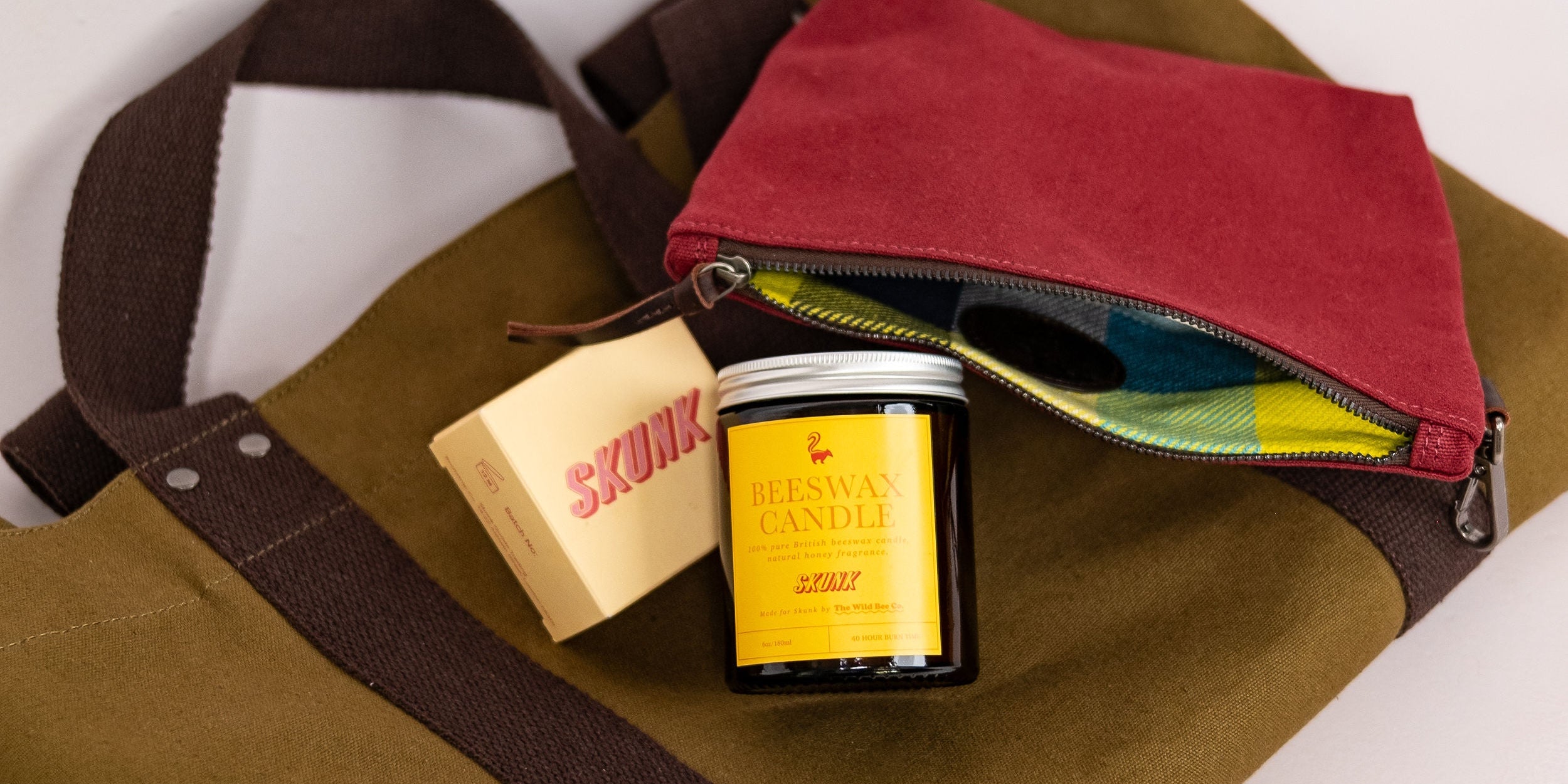 Skunk's olive green canvas tote bag with dark brown straps, styled with a burgundy zip pouch, a yellow 100% pure bees wax candle, and a bar of cold process soap